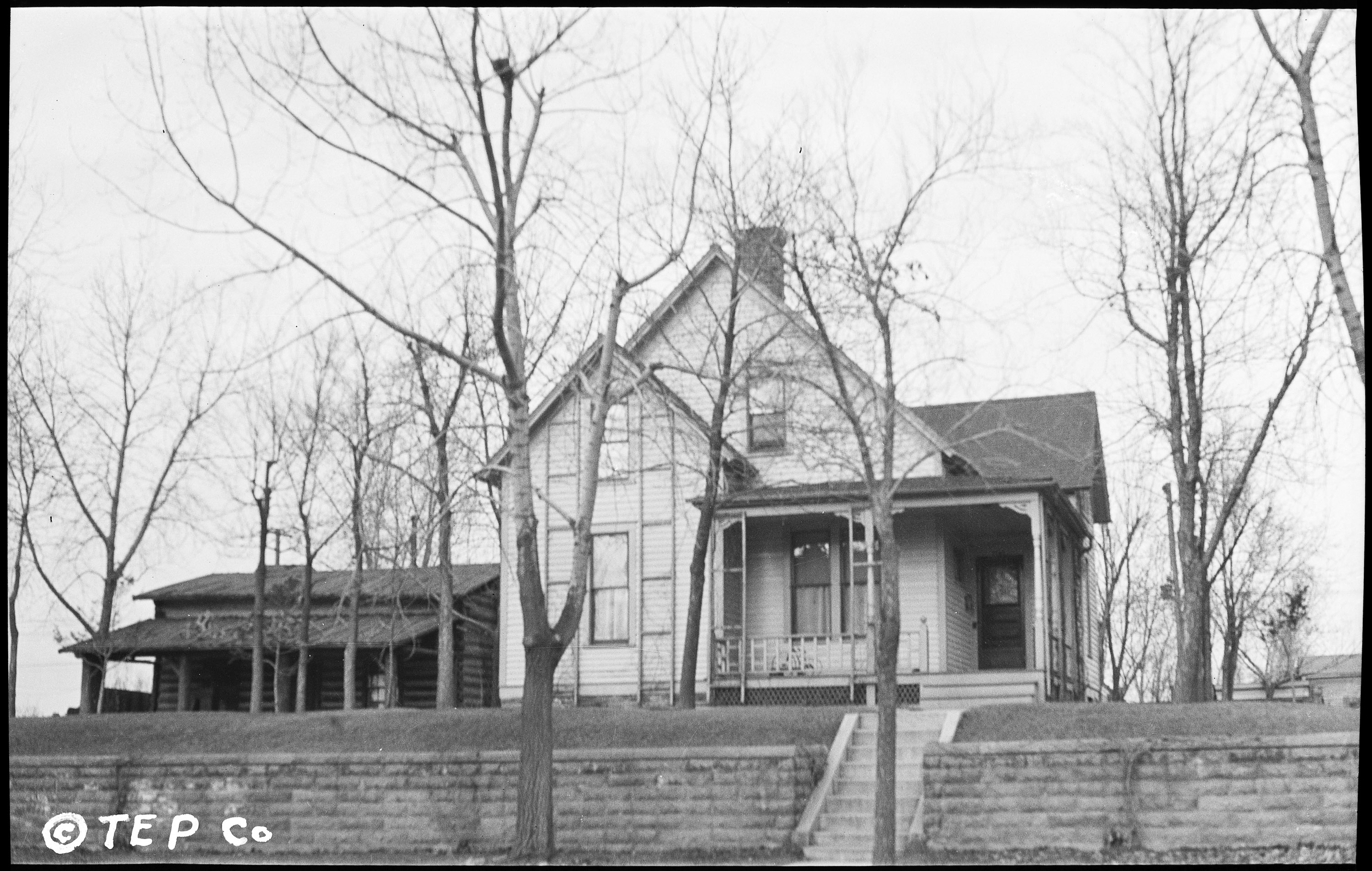 A black-and-white photograph of a two-story clapboard house with a wrap-around-porch; a log cabin off to one side.