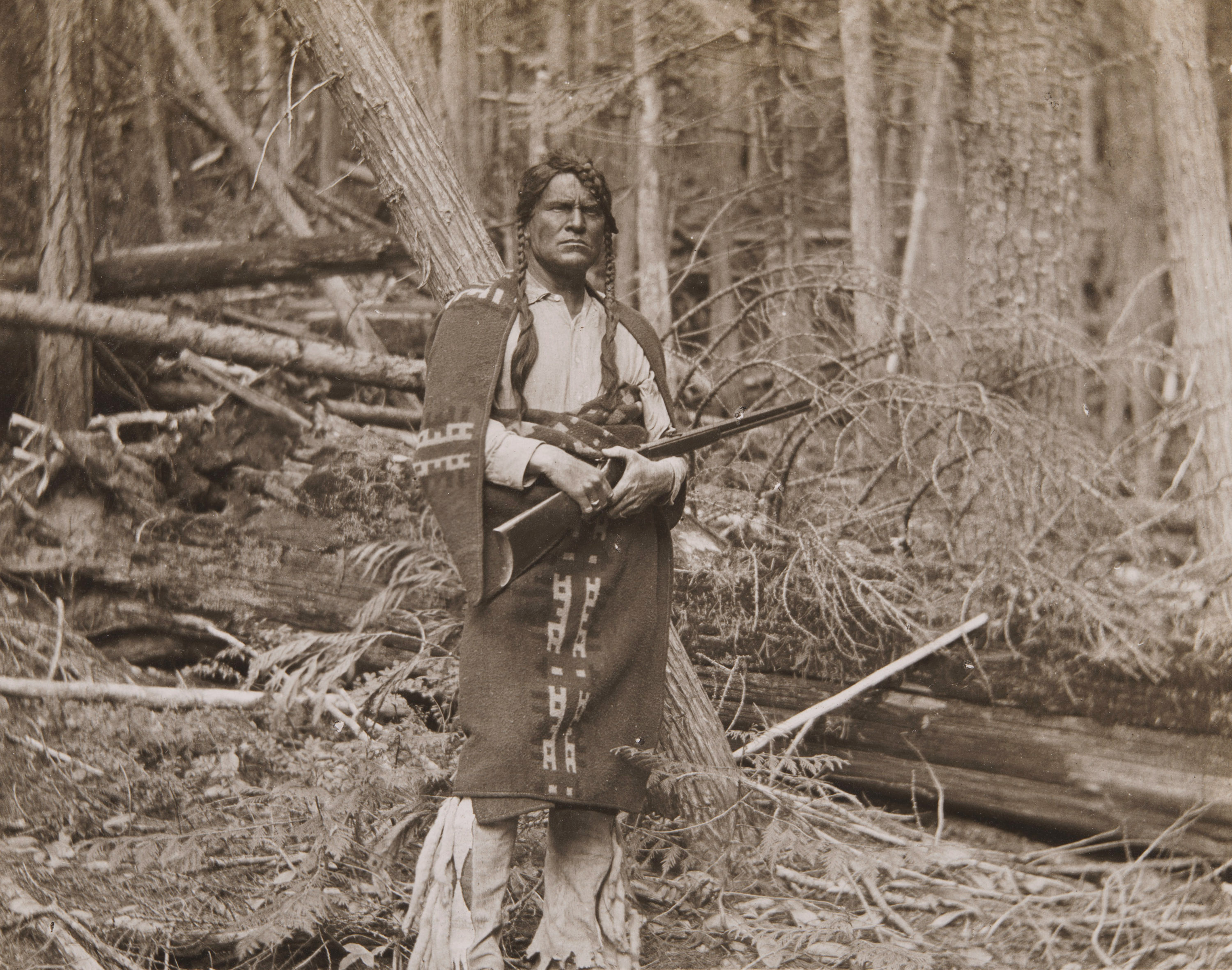 A sepia-toned photograph of a White man, his face painted to look like a Native American, wearing long braids, blankets around his shoulders, holding a rifle.