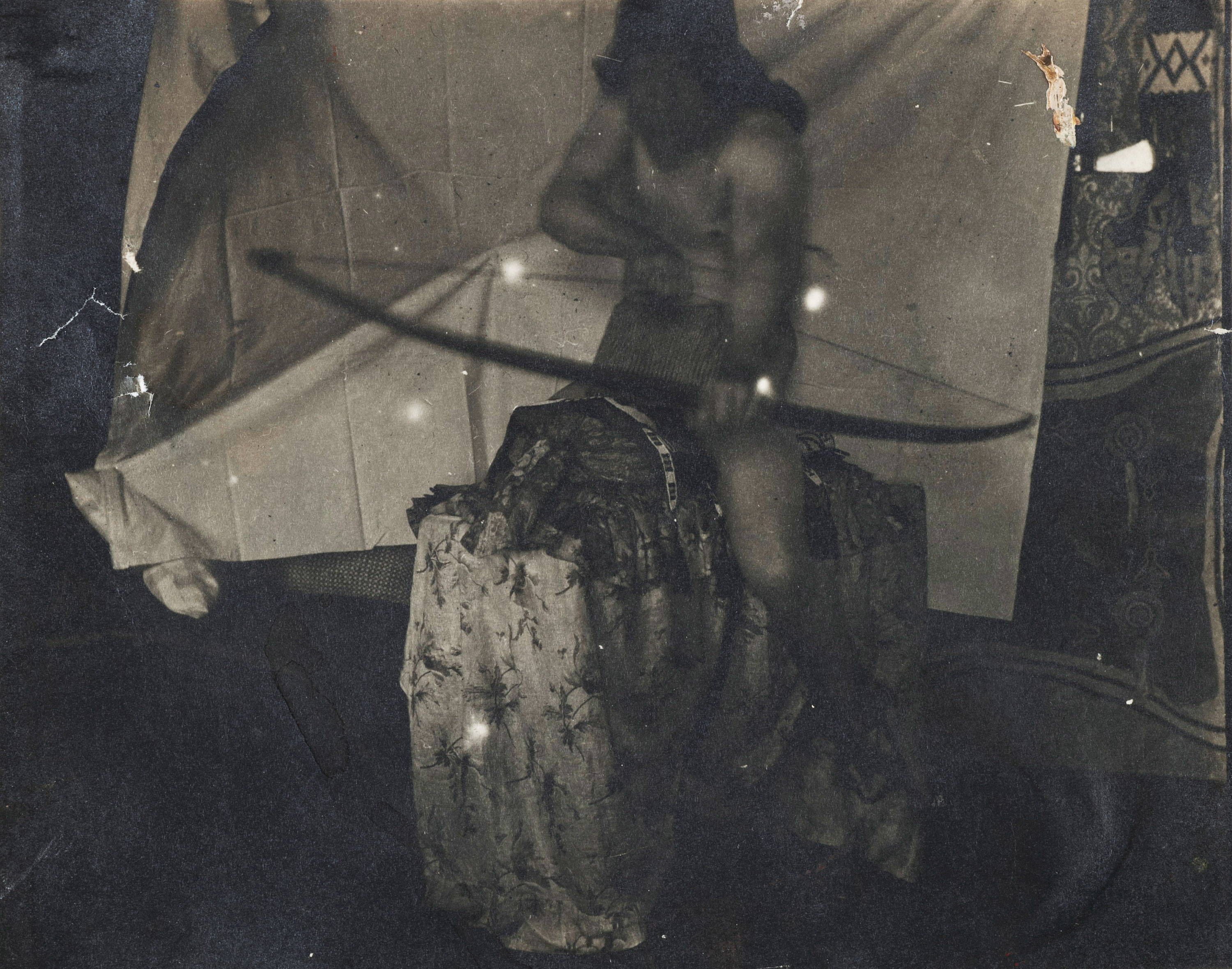 A black-and-white photograph, slightly out of focus, of a shirtless white man with a cloth over his head, pointing a bow and arrow at the camera, while sitting on a makeshift horse.