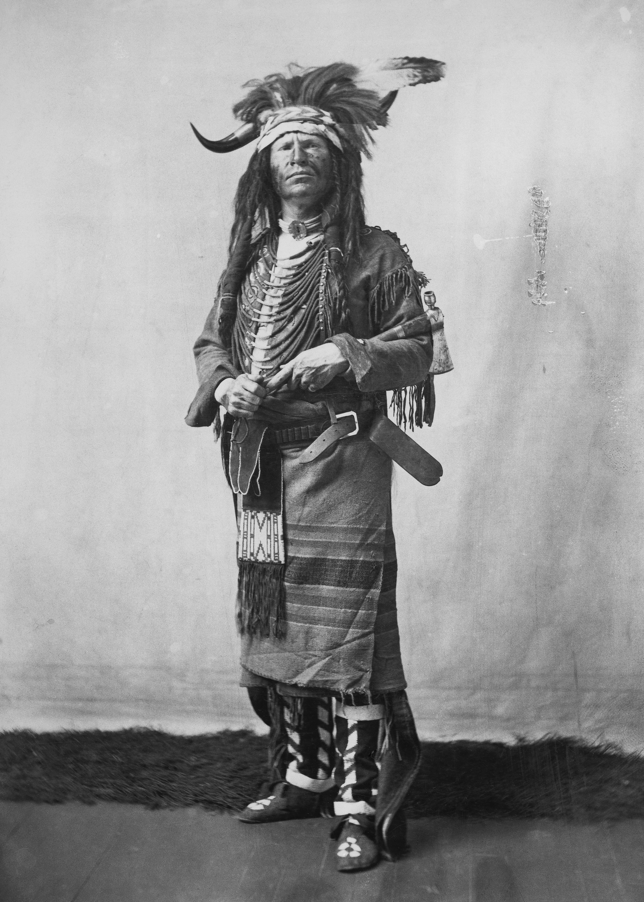 A black-and-white photograph of a White man, face painted, wearing a headdress of feathers and bison horns, a beaded necklace, fringed jacket and tunic.