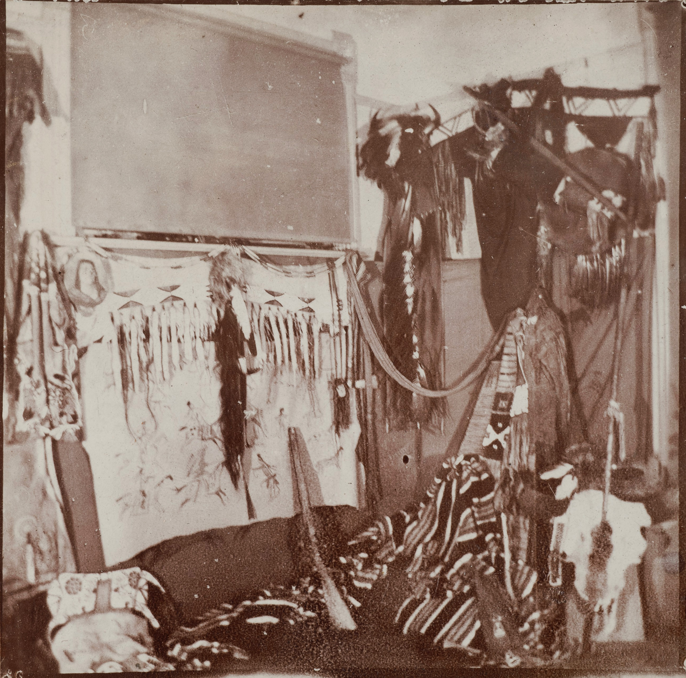 A sepia-toned photograph of the corner of a room filled with Native American blankets, clothing, and other paraphernalia.