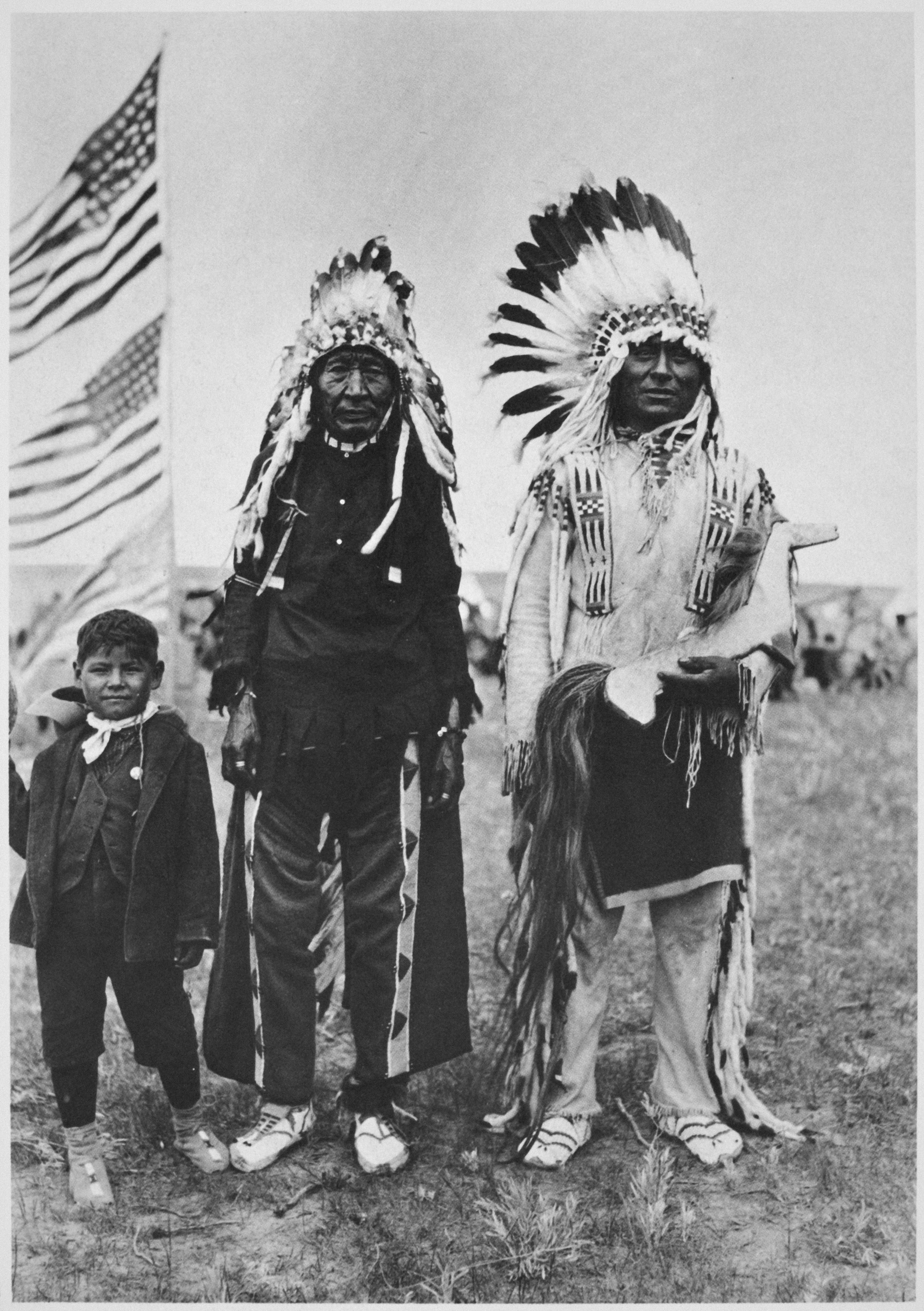 A black-and-white photograph of three Indigenous people: a child and two adults dressed in native clothing including full feathered headdresses.