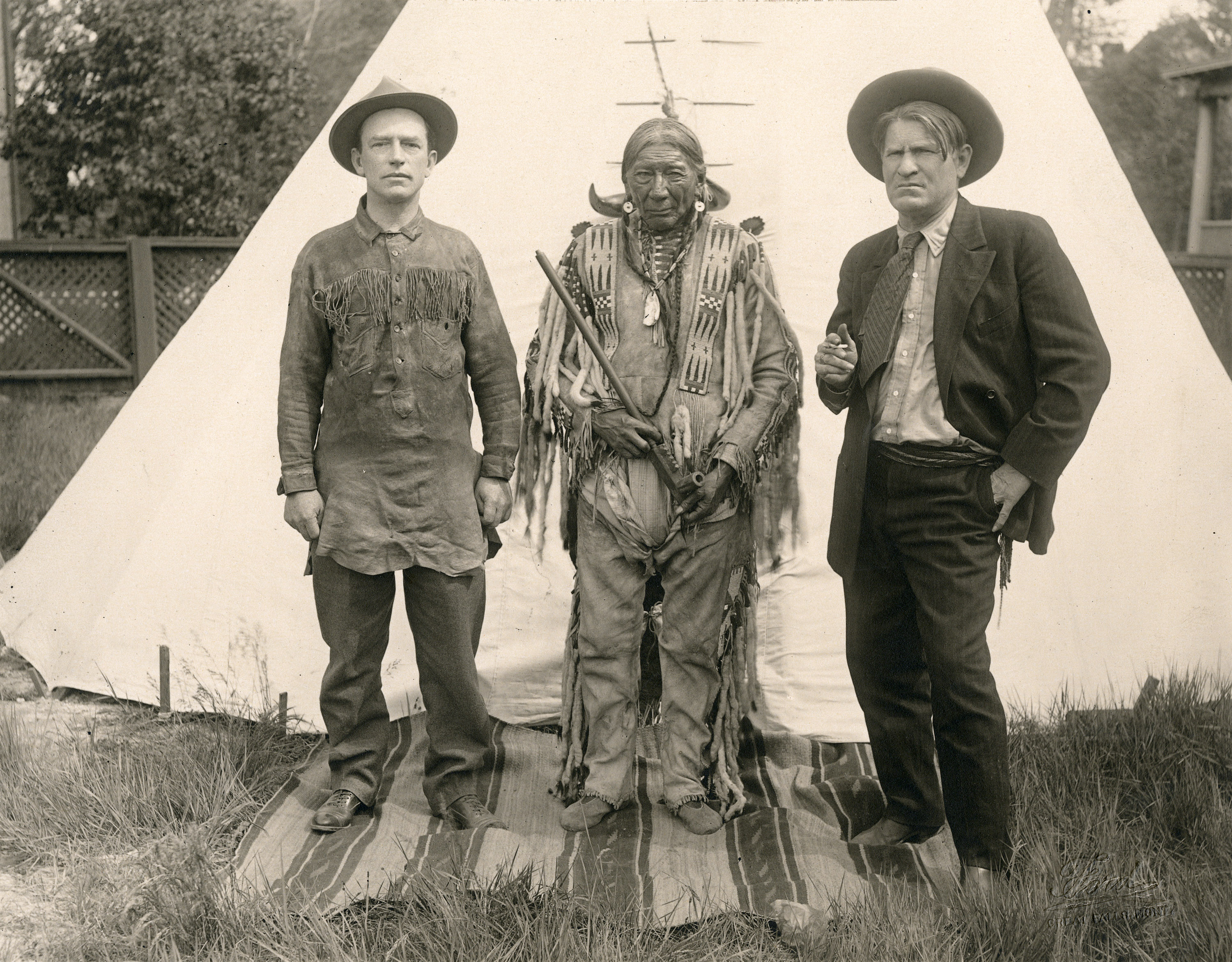 A black-and-white photograph of three men standing in front of a tipi: a White man wearing a fringed shirt, an Indigenous man wearing native clothing and holding a peace pipe, and a White man wearing a cowboy hat, jacket and tie.