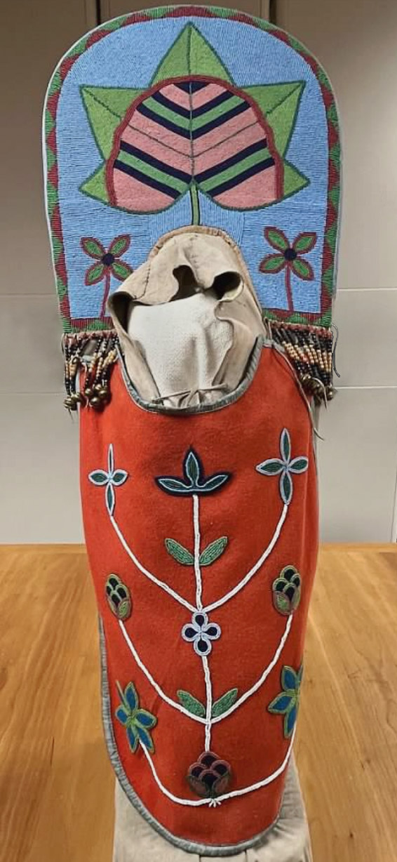 A Native American cradleboard, the back heavily beaded with a blue, green, and pink design and a red pouch with a beaded floral design.
