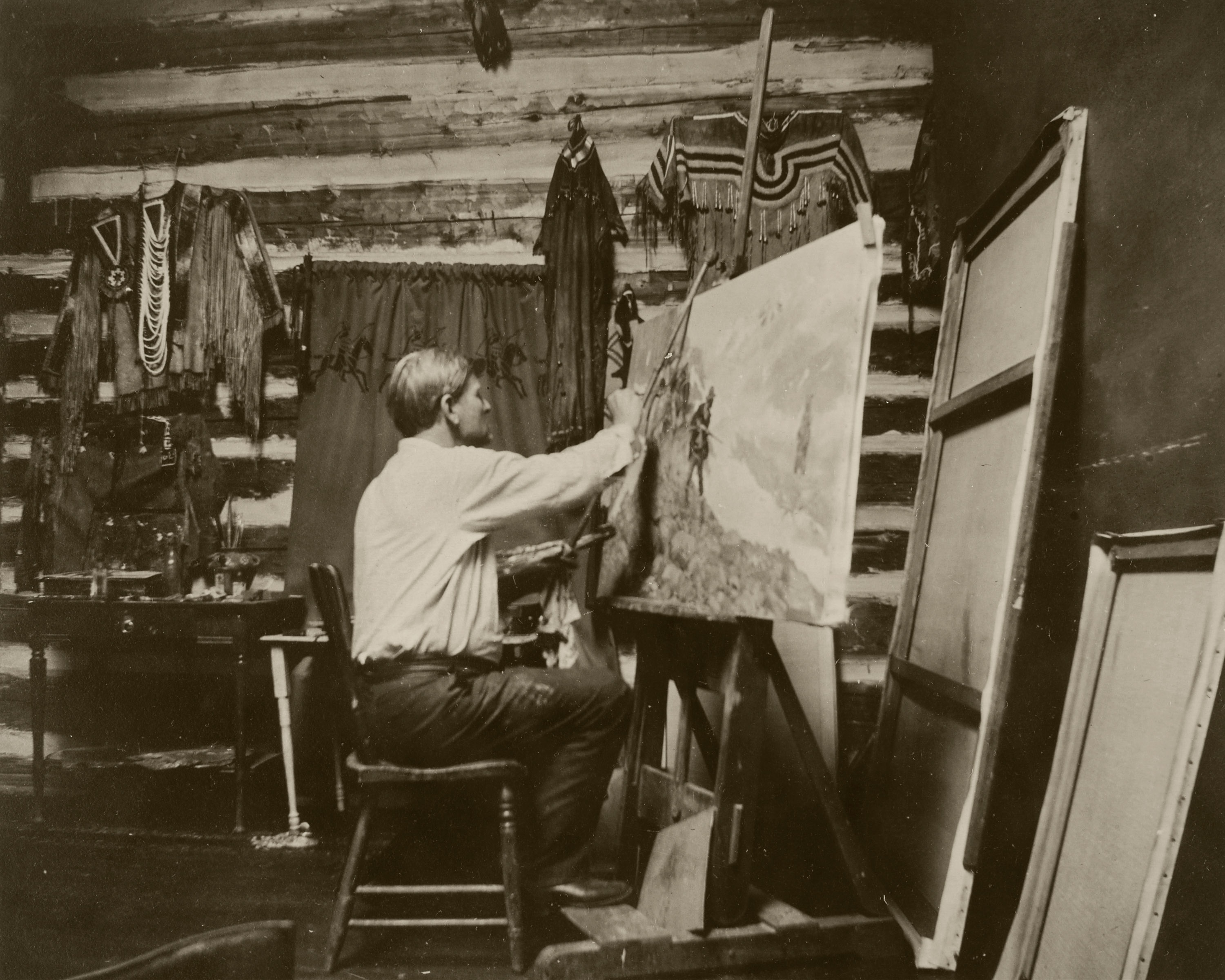 A black-and-white photograph of a White man seated in front of a easel painting on a canvas inside of what looks like a log cabin.