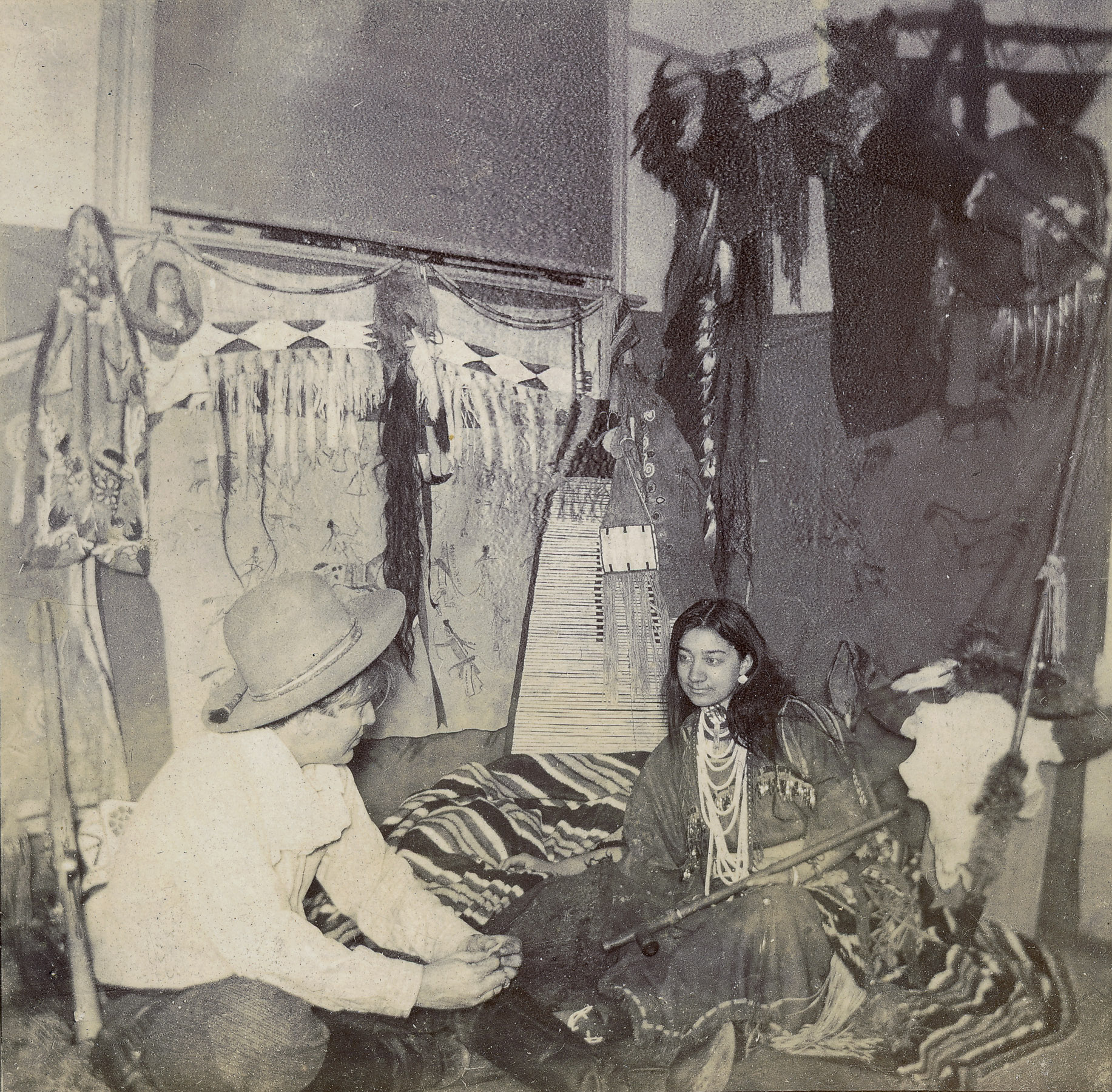 A black-and-white photograph of a White man wearing a cowboy hat seated on what looks like a sofa covered in blankets next to a woman with long dark hair wearing beaded necklaces over a long flowing dress.