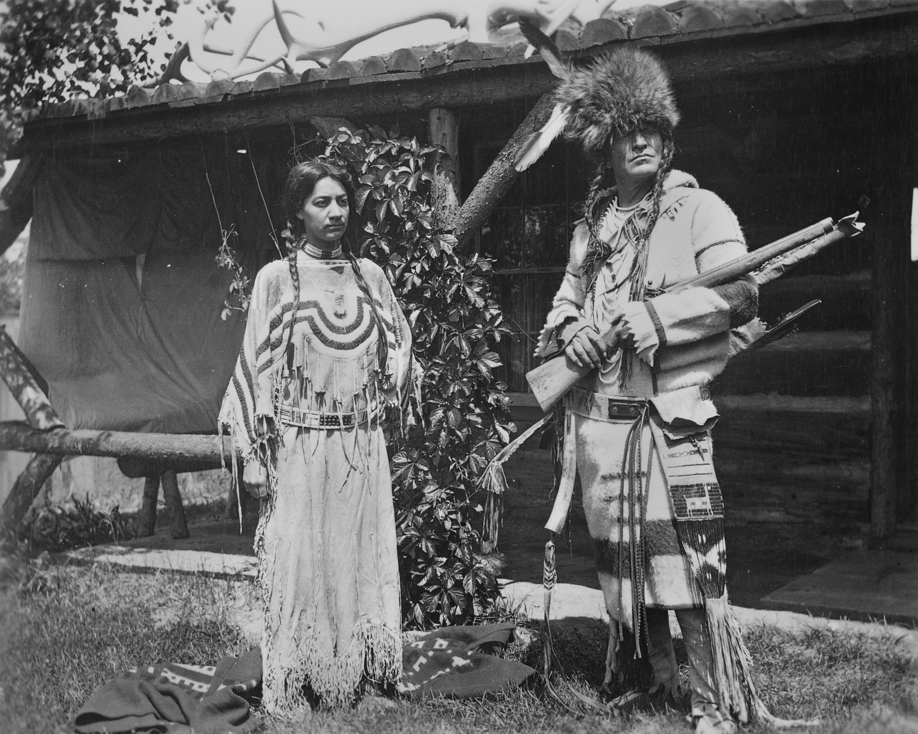 A black-and-white photograph of a woman and man posing outside of a building both dressedin Native American clothing.