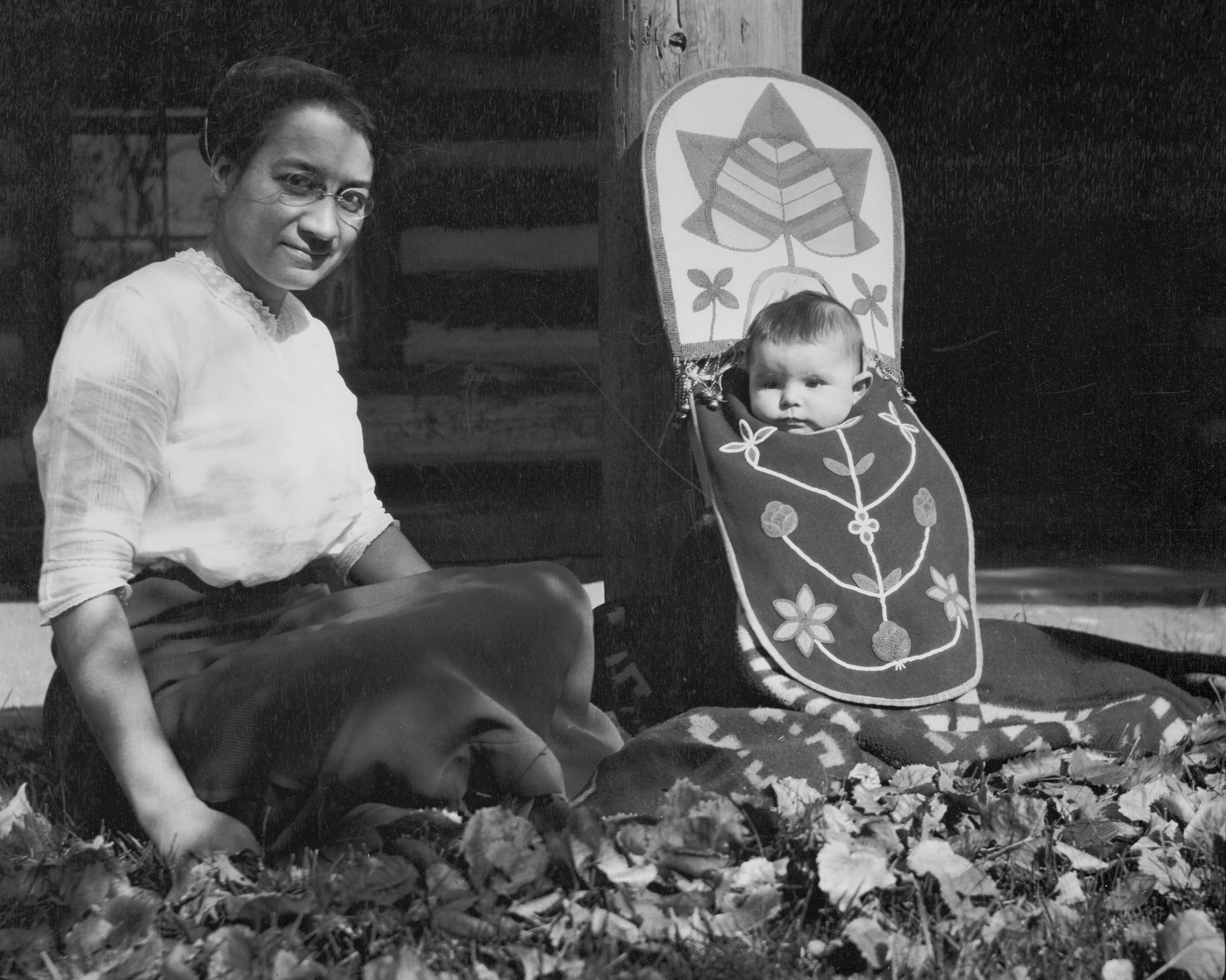 A black-and-white photograph of a woman, face in shadow, seated on the ground next to a baby in a beaded cradleboard that is leaning on a post.