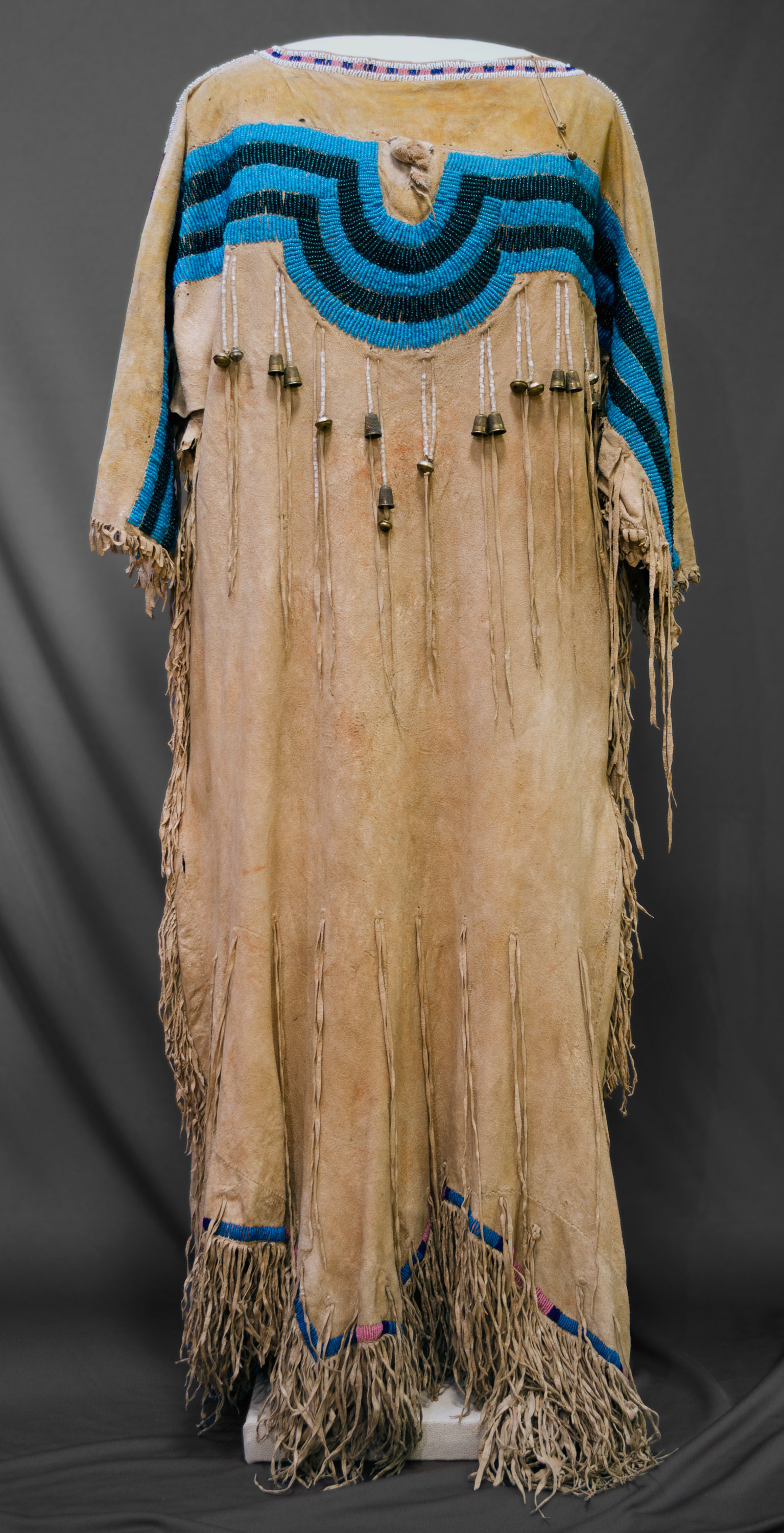 An animal hide dress, fringe along the sleeves, sides, and bottom, a blue and black beaded design along the top with what looks like metal bells hanging beneath the design.