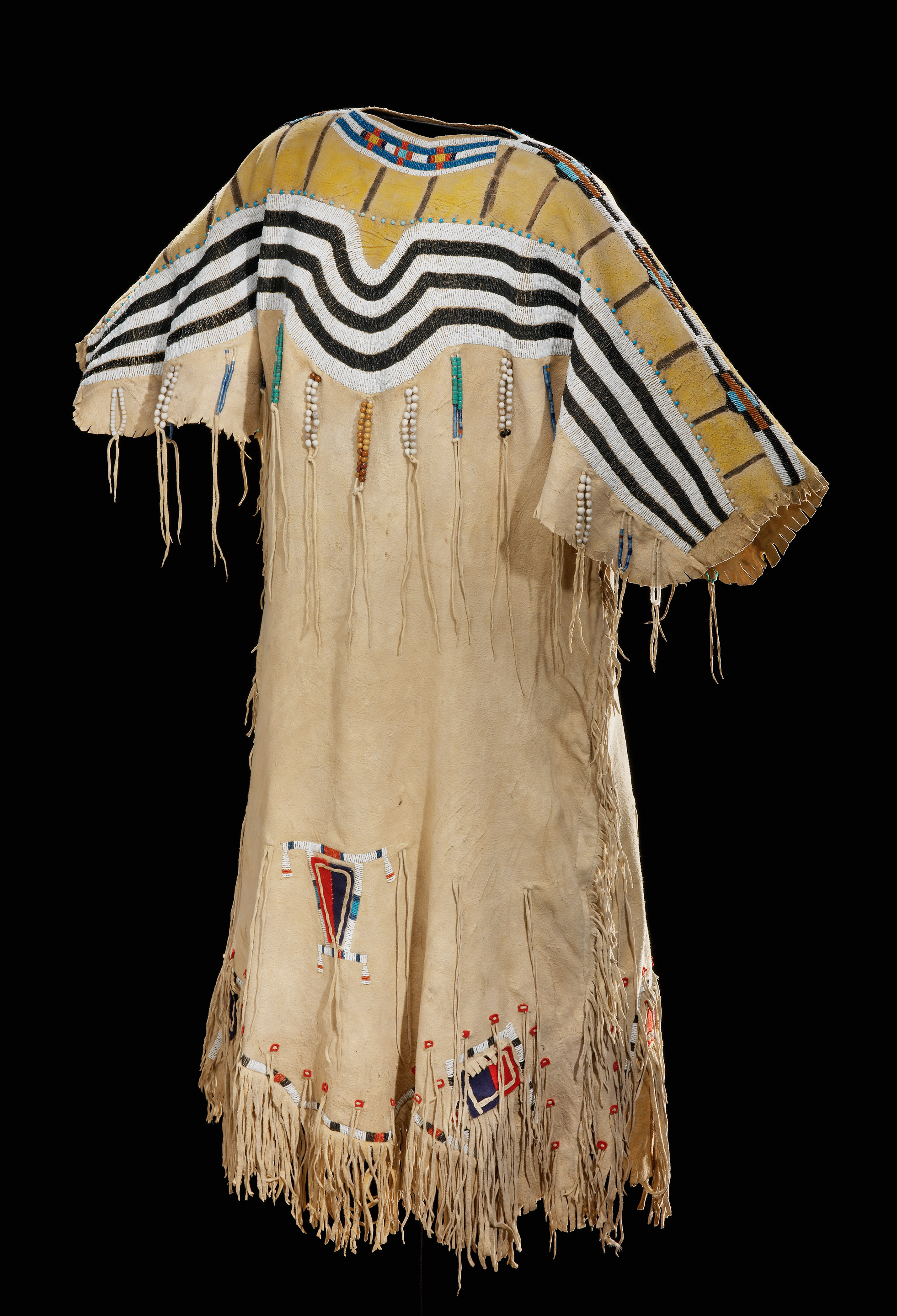 An animal hide dress, fringe along the sleeves, sides, and bottom, a black and white beaded design along the top with beaded fringe hanging beneath the design.