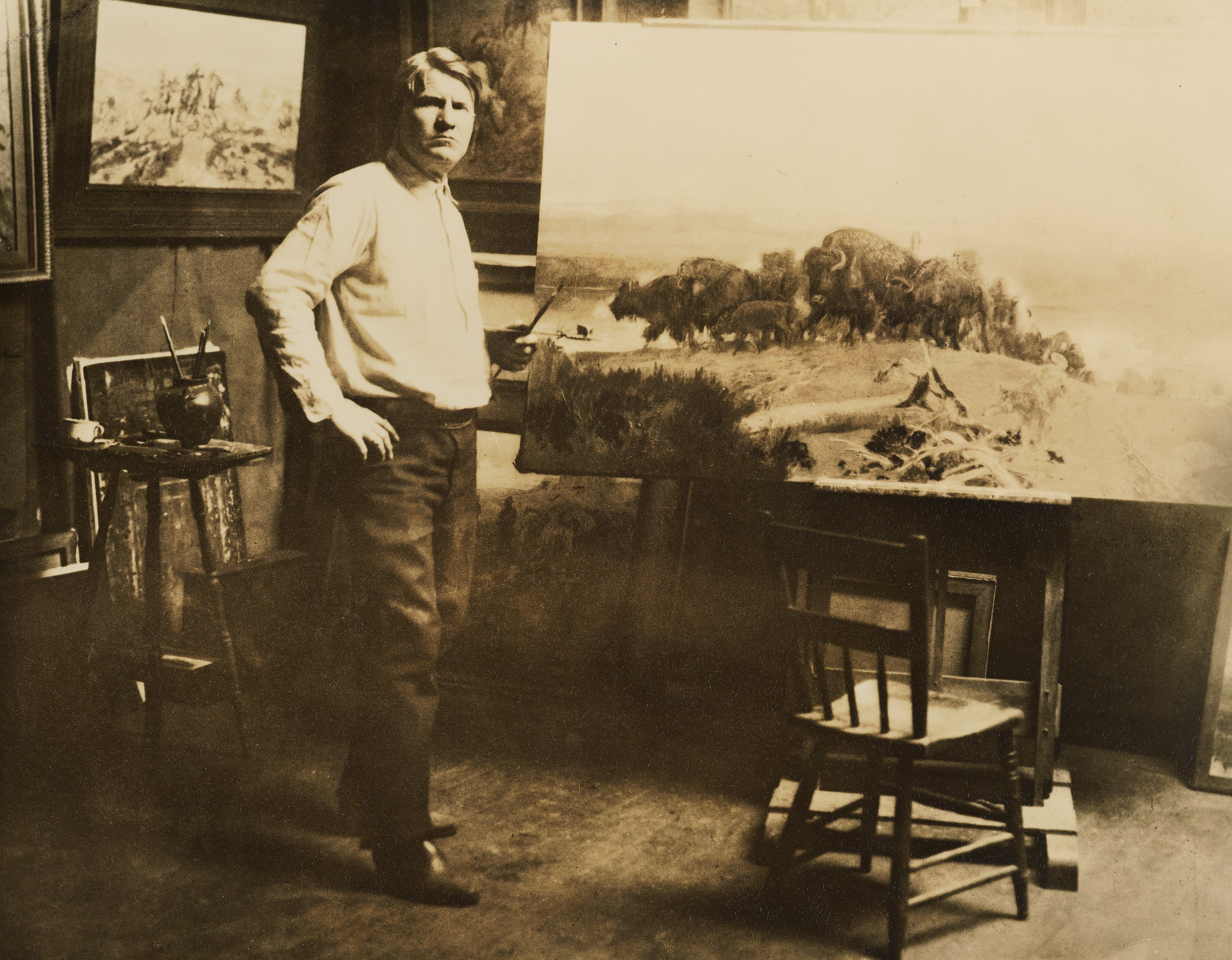 A sepia-toned photograph of a White man posing next to a large canvas that depicts a herd of bison.